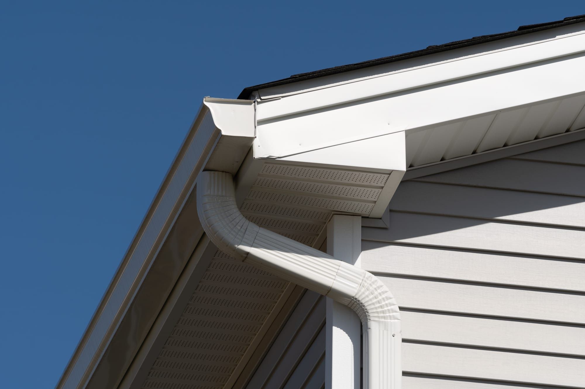 White seamless gutter, downspout, and vented soffit along a home's roofline against a clear blue sky